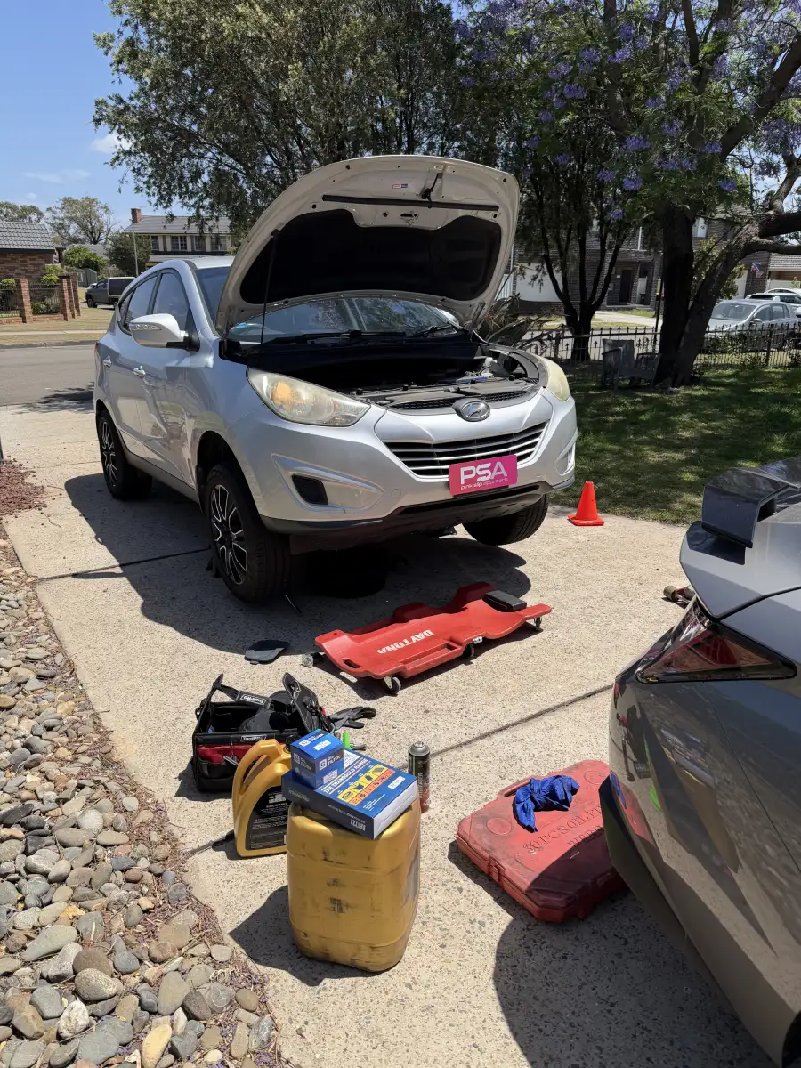 A silver Hyundai with its bonnet up in a driveway, tools and oil laid out beside it
